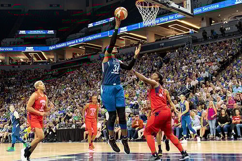 A basketball player in a blue uniform jumps toward a basket with a ball in hand. Three players wearing red uniforms surround her.